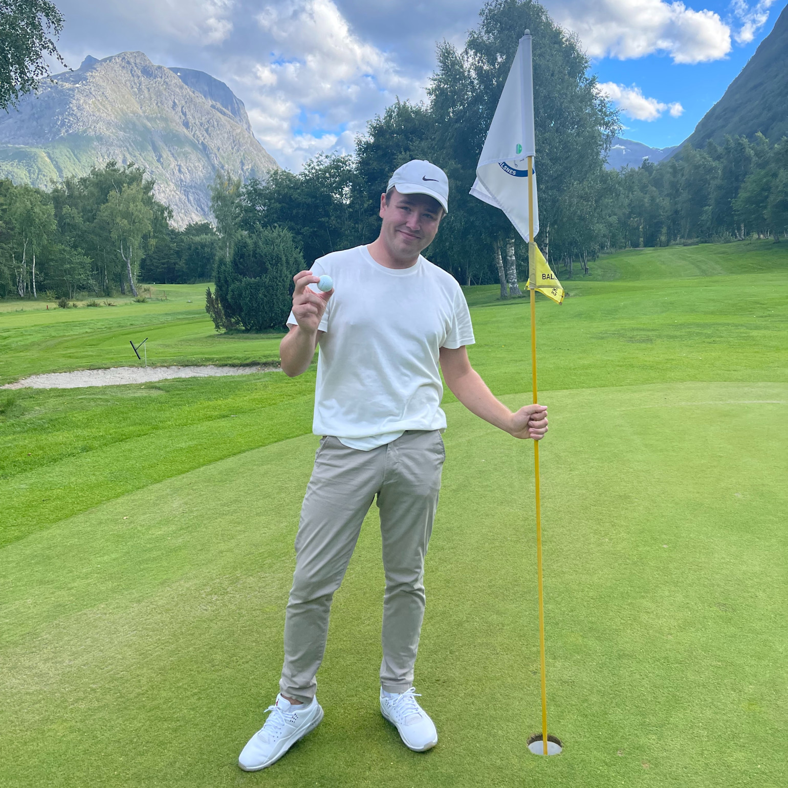 Mand i hvid t-shirt og kasket holder en golfbold og en flagstang på en golfgreen med bjerge og træer i baggrunden.