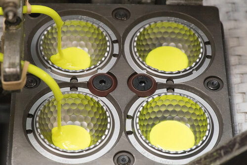 Fyra formar fylls med gul vätska genom slangar under tillverkning av golfbollar i en fabriksmiljö.