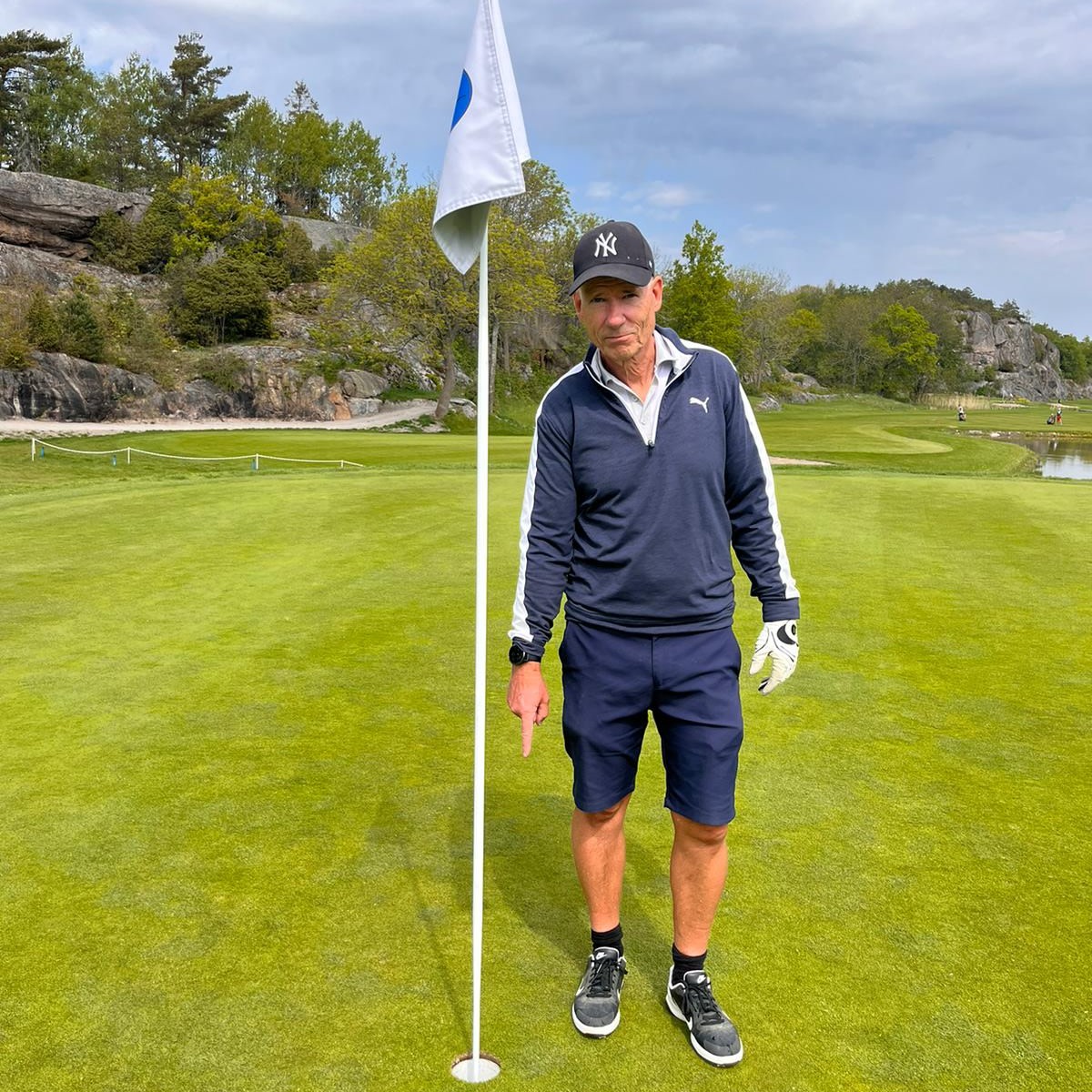 Mand i golfbeklædning peger mod et golfhul ved siden af en flagstang på en grøn golfbane med træer og klipper i baggrunden.