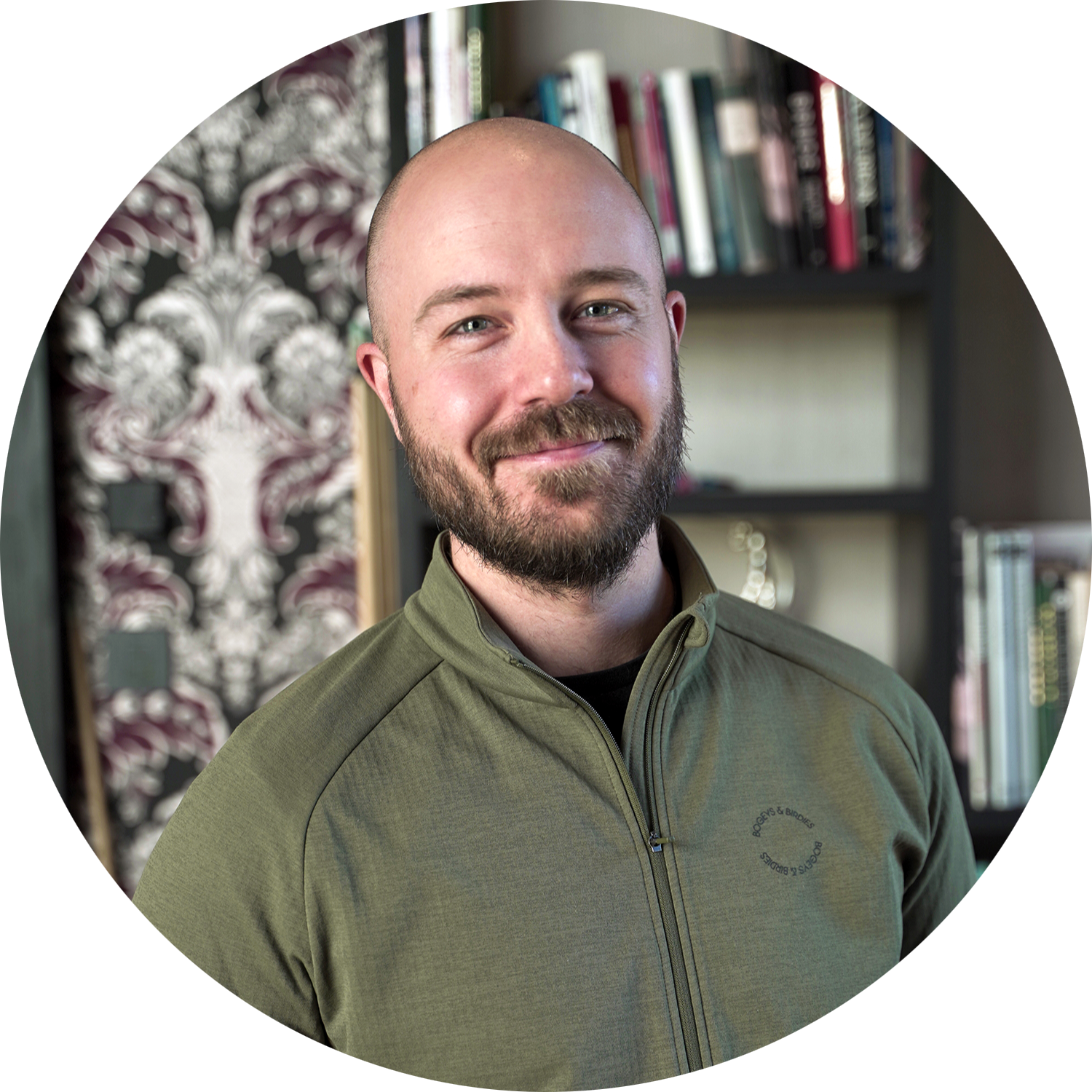 Skäggig man med rakat huvud ler och står framför en bokhylla med böcker och mönstrad tapet i bakgrunden.