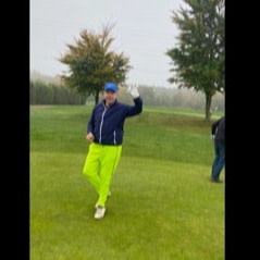 En person iført en blå jakke og neongule bukser vinker og smiler på en golfbane med grønt græs og træer i baggrunden.