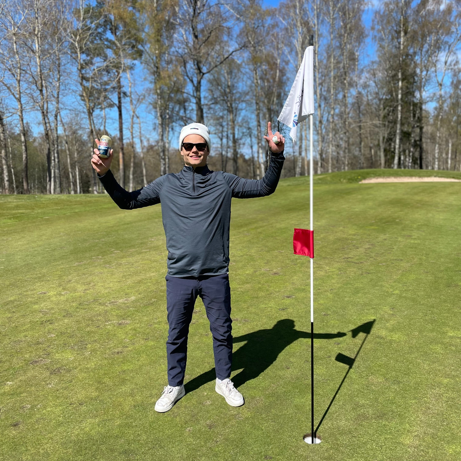 Man i solglasögon och mössa står bredvid en golfhålsflagga på green, håller upp en golfboll och en burk, omgiven av träd.