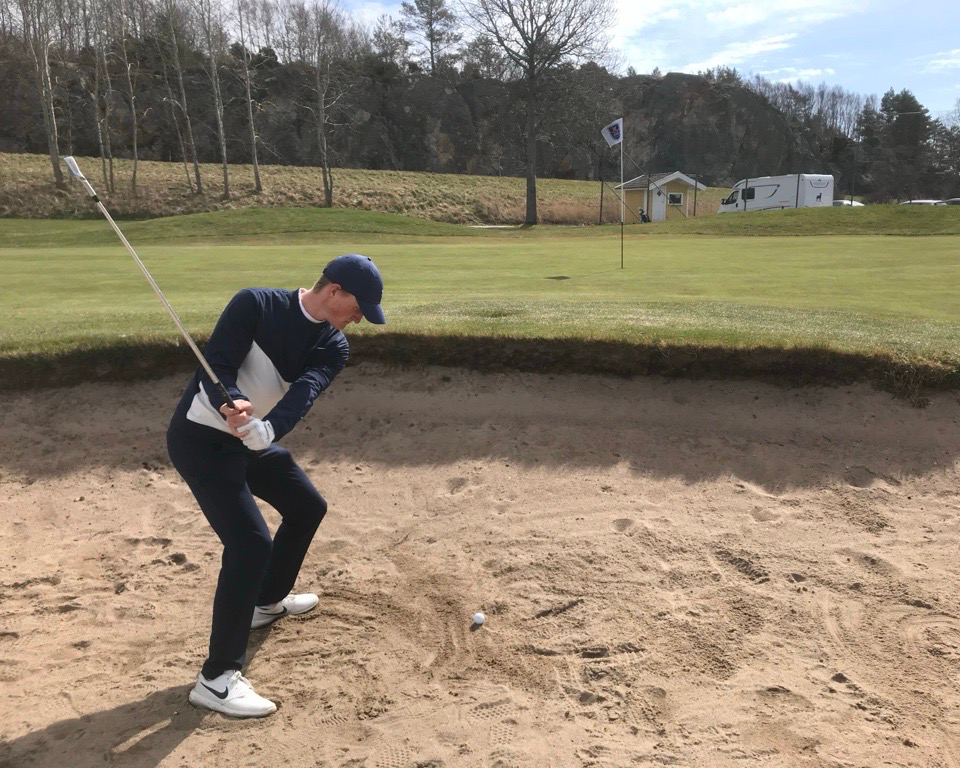 Golfspelare svingar en klubba för att slå en boll ur en sandbunker mot greenen på en golfbana.