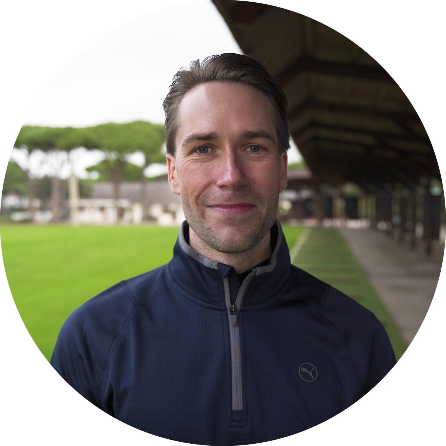 Man ler mot kameran, står utomhus på en grön gräsmatta med träd och byggnader i bakgrunden.