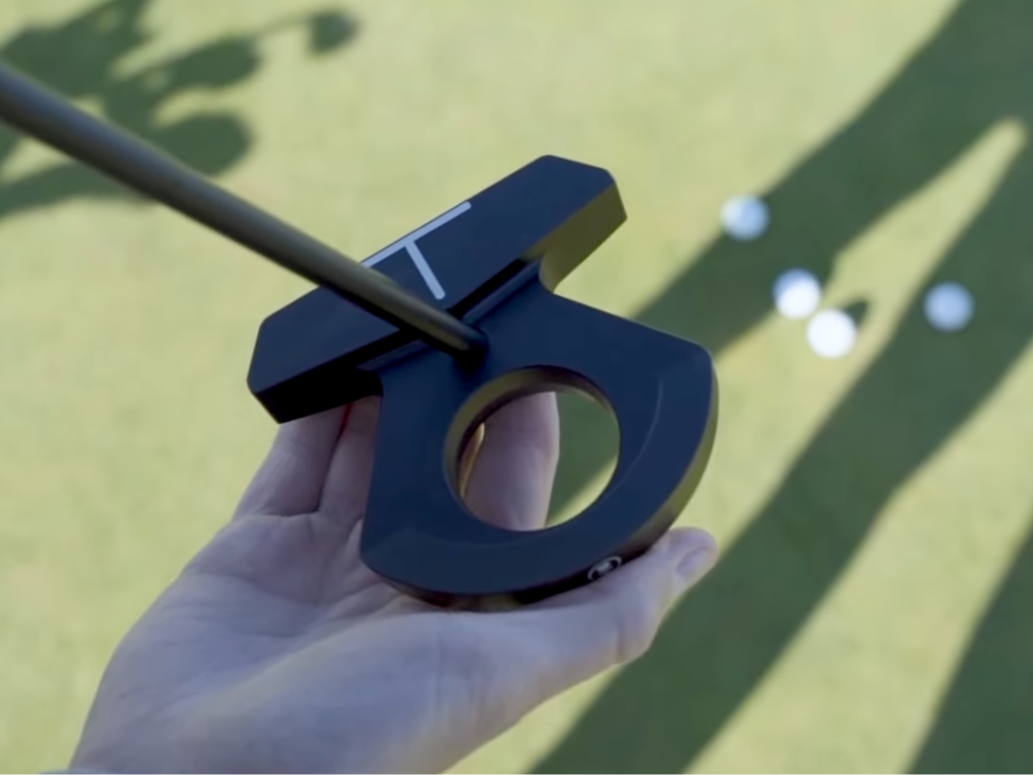 Eine Hand hält einen modernen Golf-Putter mit einer kreisförmigen Öffnung, während Golfbälle und deren Schatten im Gras im Hintergrund zu sehen sind.
