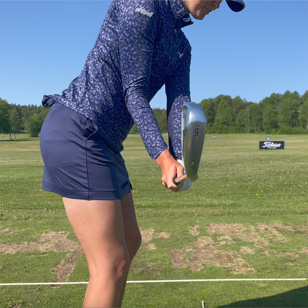 Golfspelare i blå tröja och kjol svingar en järnnia på en driving range med gräs och träd i bakgrunden.