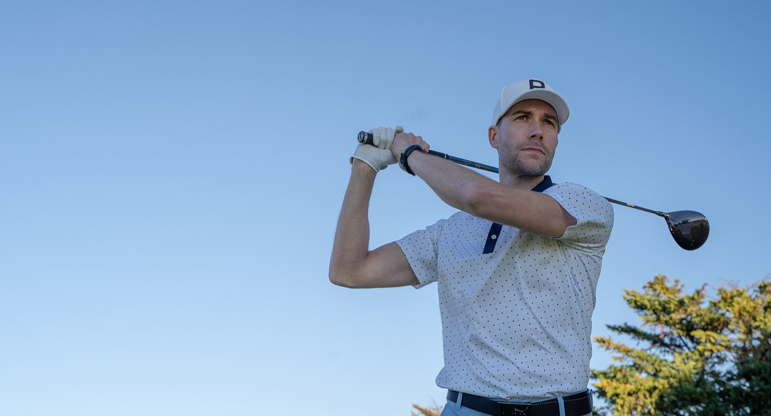 Mies, joka heilauttaa golfmailaa golfkentällä, pukeutuneena valkoiseen lippikseen ja pilkulliseen pikeepaitaan kirkkaan sinisen taivaan alla.