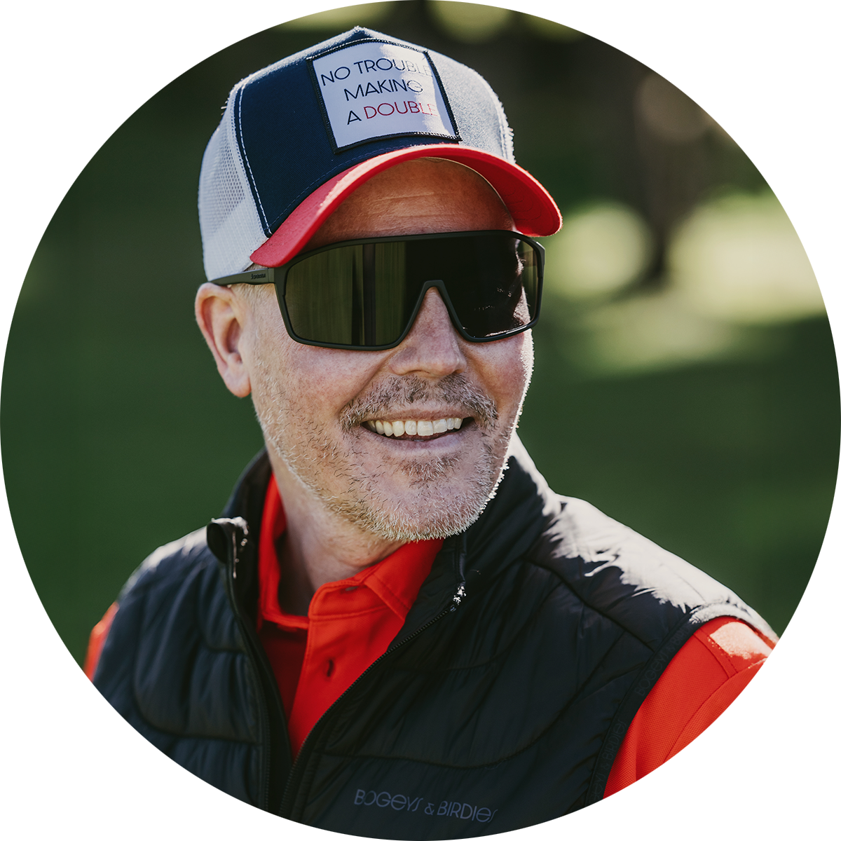 Leende man med solglasögon och keps med texten "NO TROUBLE MAKING A DOUBLE", utomhus på en grön gräsmatta.