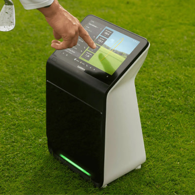 A Garmin device on a stand is positioned on a golf course next to a smartphone displaying golf data on its screen.