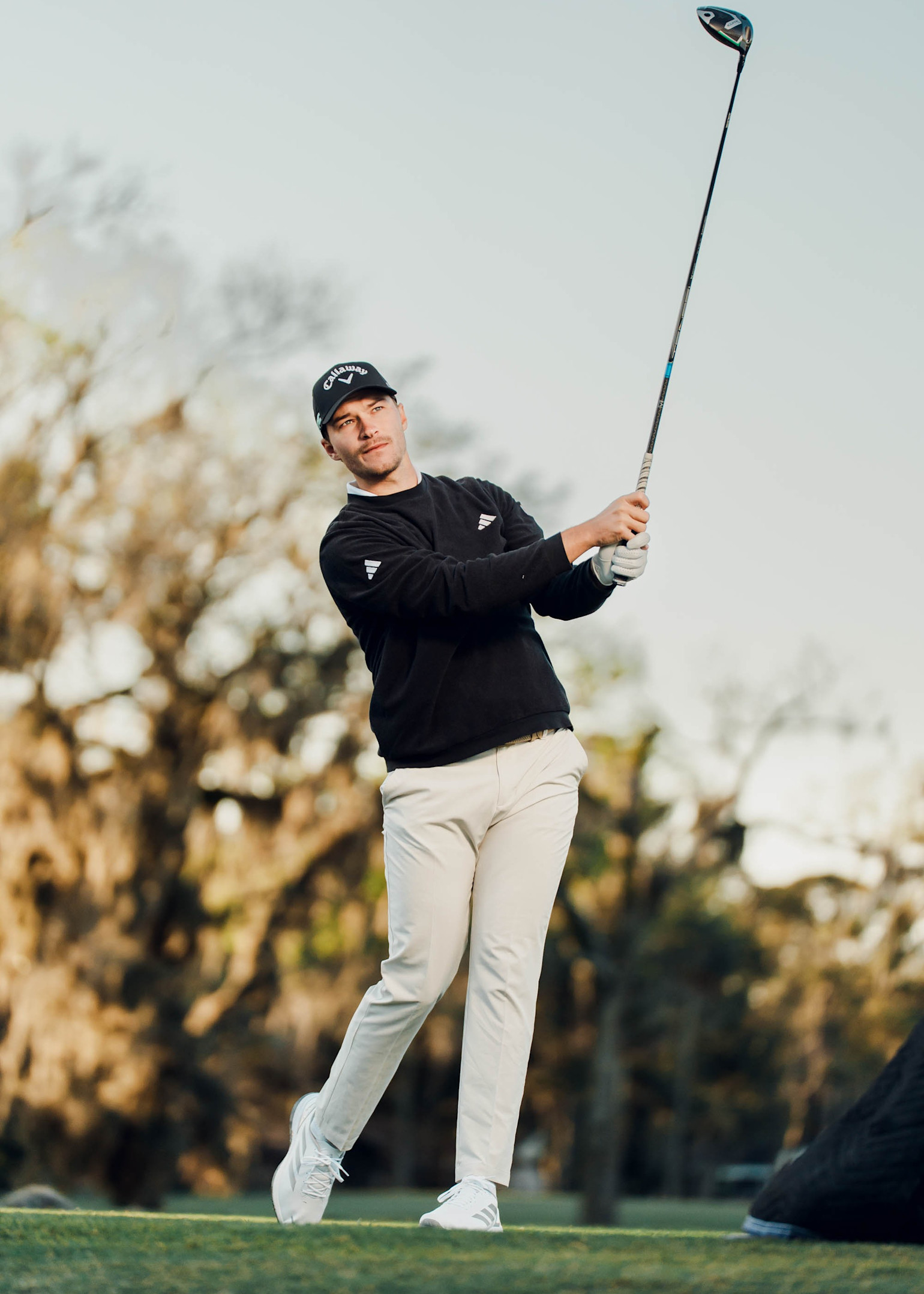 Ein Mann, der auf einem Golfplatz einen Golfschläger schwingt, gekleidet in einem schwarzen Pullover, einer hellen Hose und einer Kappe, mit Bäumen im Hintergrund.