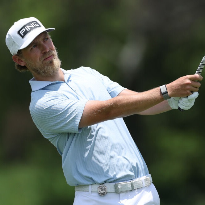 Mann i lyseblå pikéskjorte og hvit caps med teksten "PING" svinger en golfkølle utendørs på en grønn golfbane.