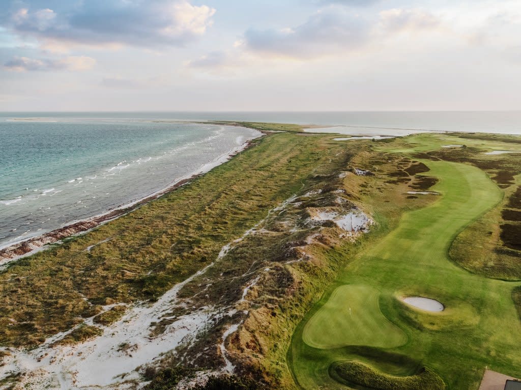 Golfbana med gröna fairways och bunkrar löper längs en kustlinje med sanddyner och hav under molnig himmel.