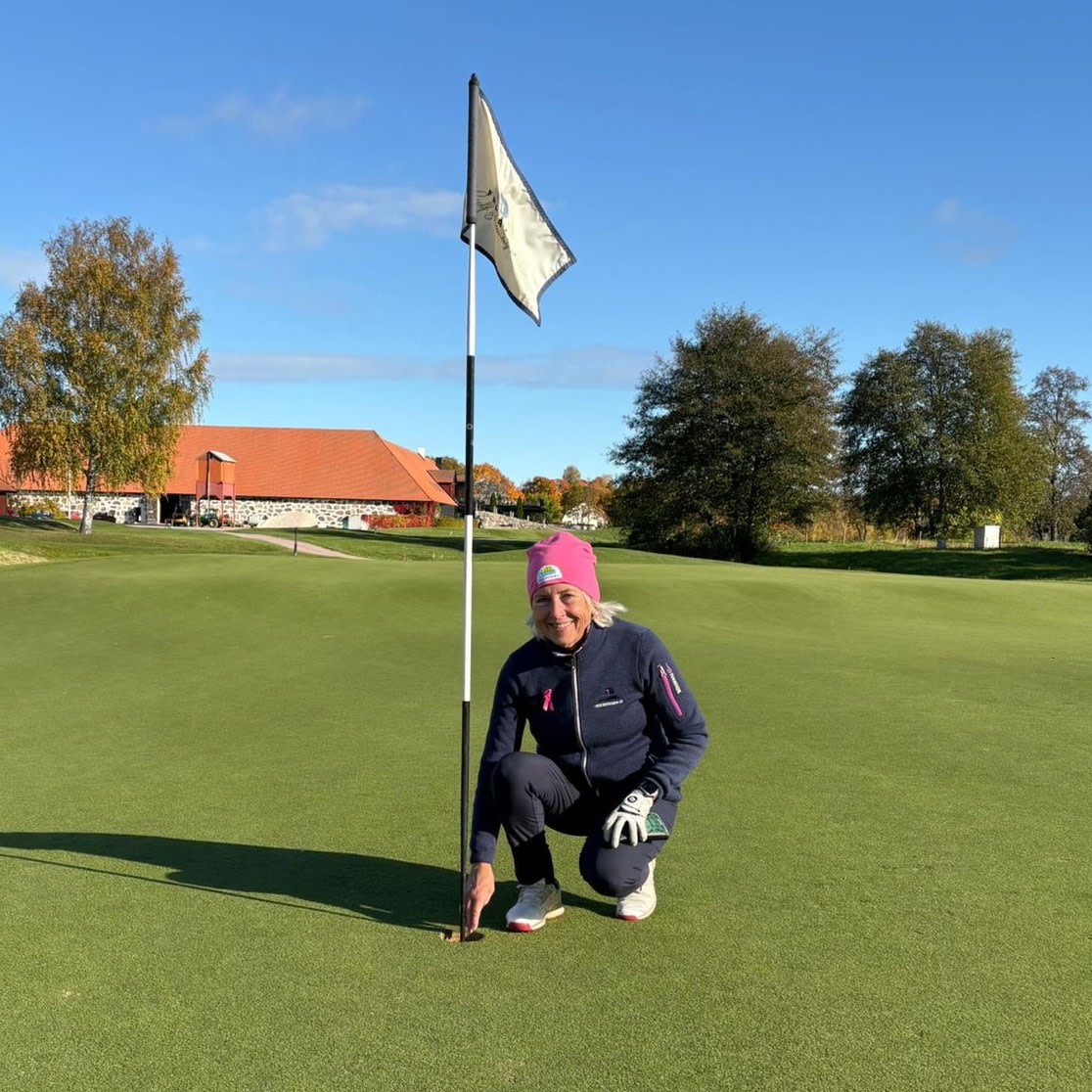 Kvinne i rosa lue huker seg ved golfhull og flaggstang på en golfbane, med klubbhus og trær i bakgrunnen.