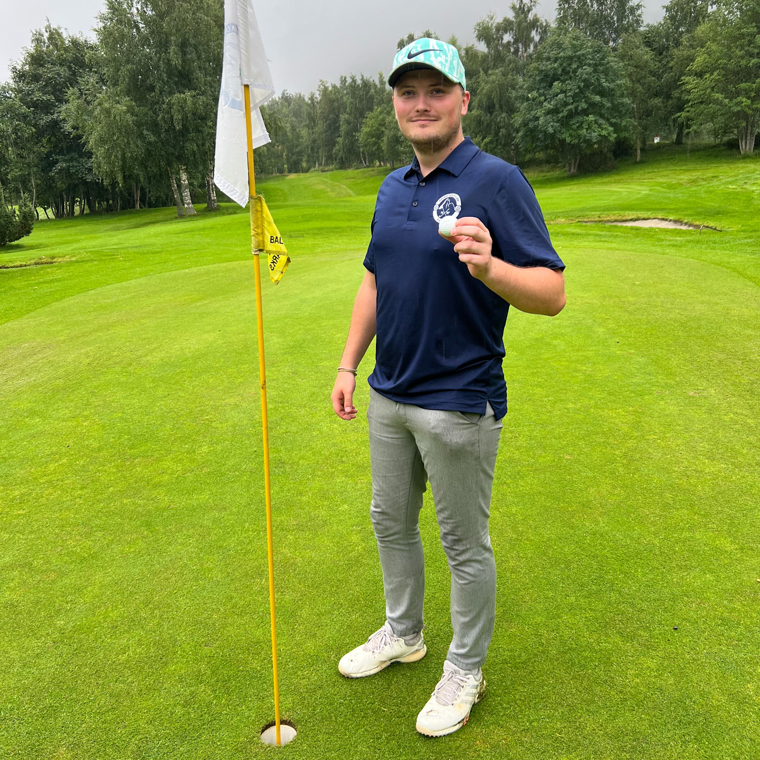 Mand iført blå polo og kasket holder en golfbold op ved siden af et hul på en golfbane med grøn natur i baggrunden.
