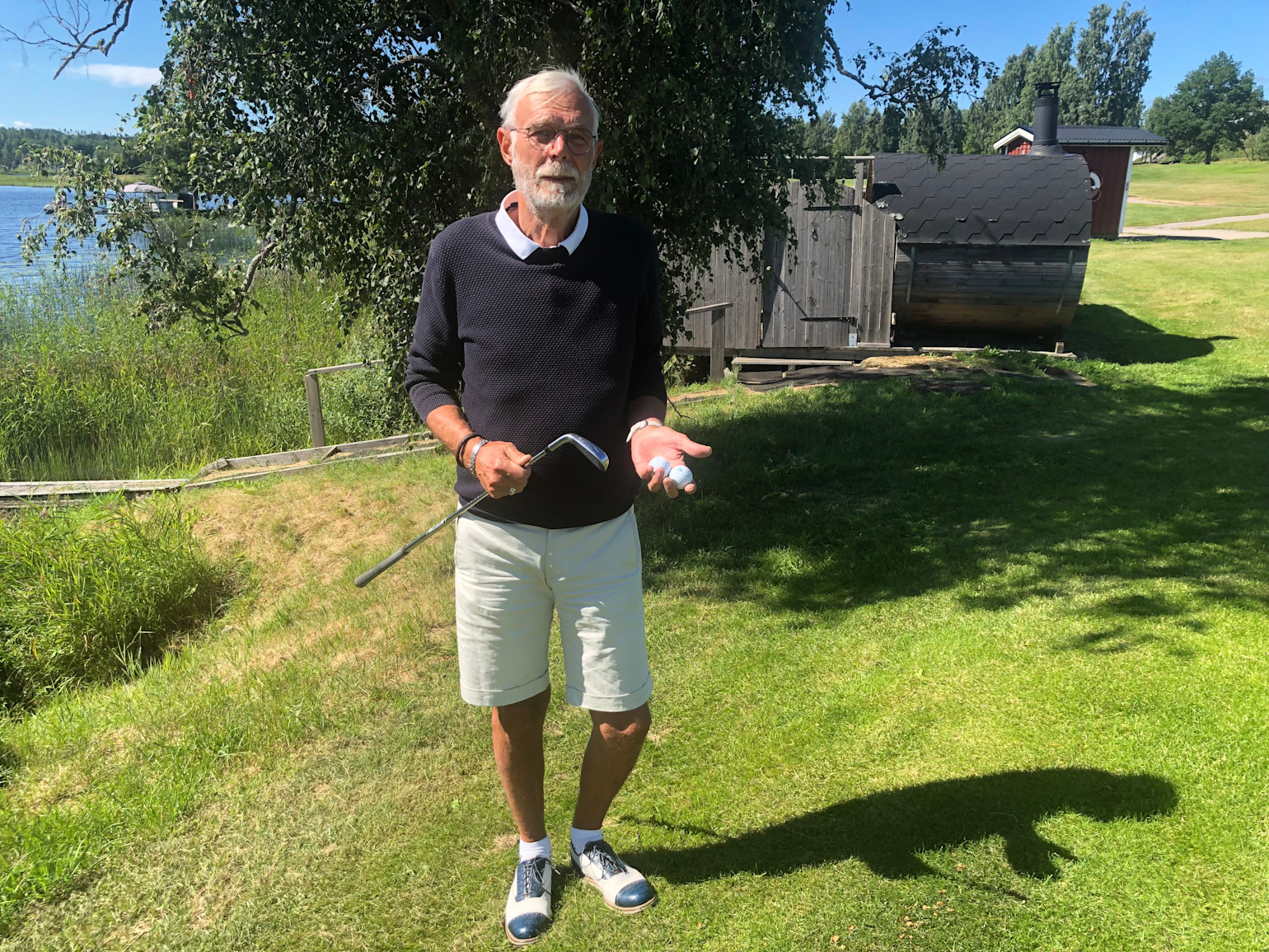 Ældre mand holder en golfkølle og to golfbolde i hånden, står på græs nær en sø og en sauna i solskinsvejr.