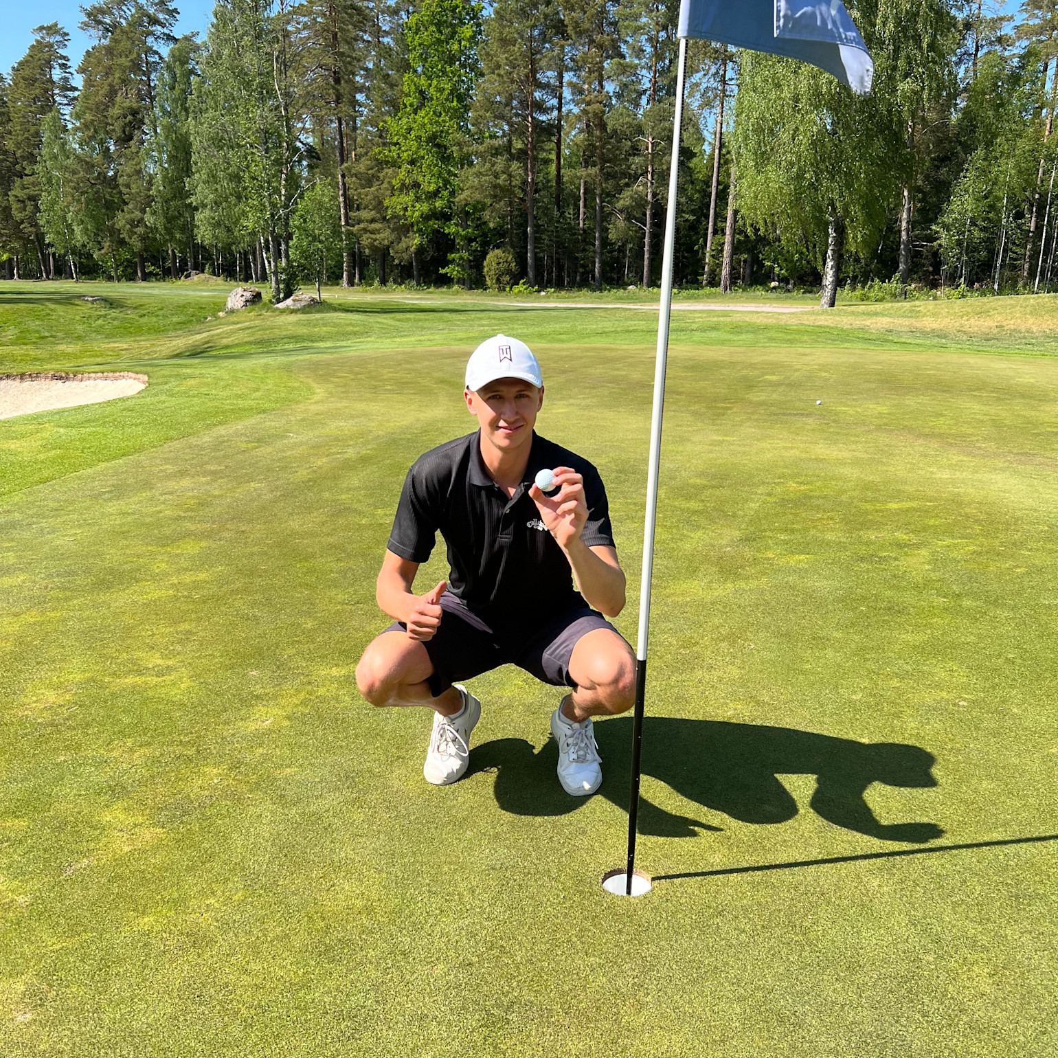 Ung mand i sort poloshirt og hvid kasket sidder på hug ved et golfhul, holder en golfbold op og giver tommelfingeren op på en solrig green.