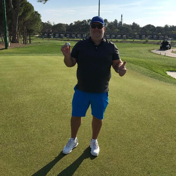 Mand i blå shorts og sort trøje holder en golfbold op og giver tommelfingeren op på en golfbane i solskin.