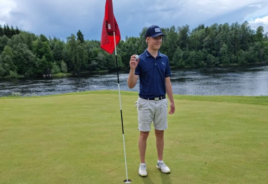 Ung mand i golfbeklædning holder en golfbold op ved siden af flagstangen på greenen, med en sø og skov i baggrunden.