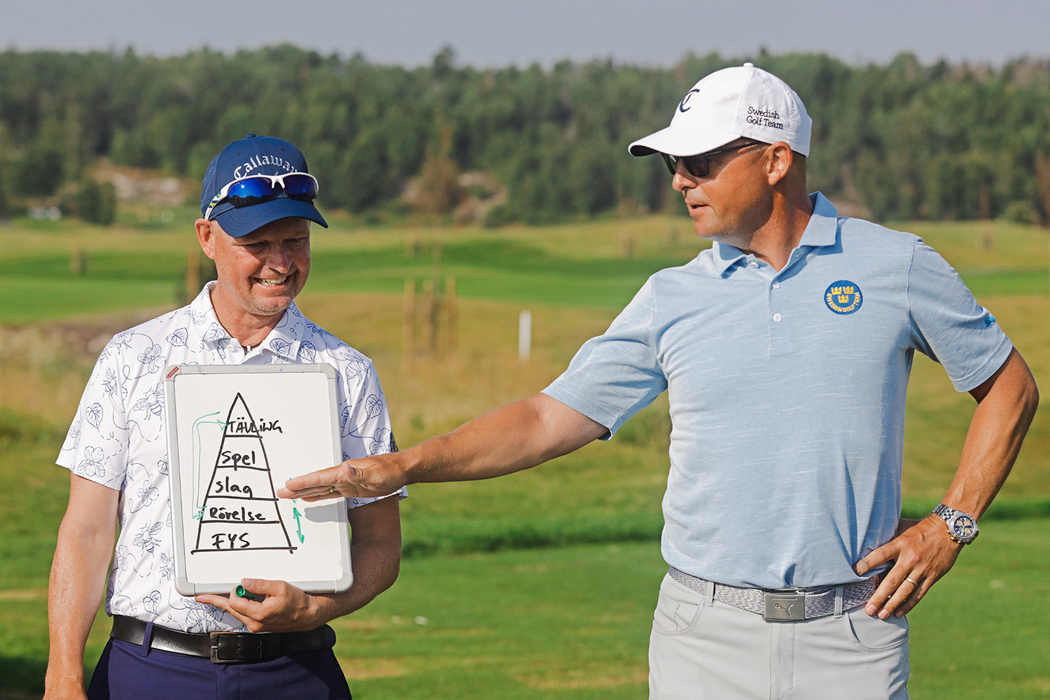 Två män står på en golfbana, en håller en whiteboard med en pyramid där det står "Tävling, Spel, Slag, Rörelse, FYS", den andra pekar på tavlan.