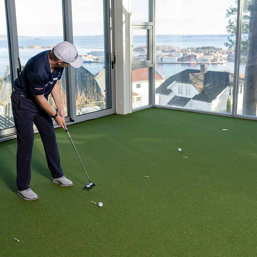 En man puttar en golfboll på en konstgräsmatta inomhus med stora fönster och utsikt över havet och hus.