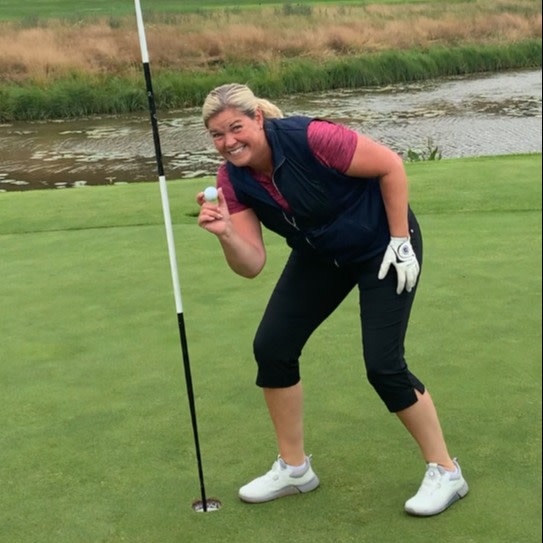 Kvinde smiler og holder en golfbold op ved siden af et golfhul på greenen, med en golfbane og vand i baggrunden.