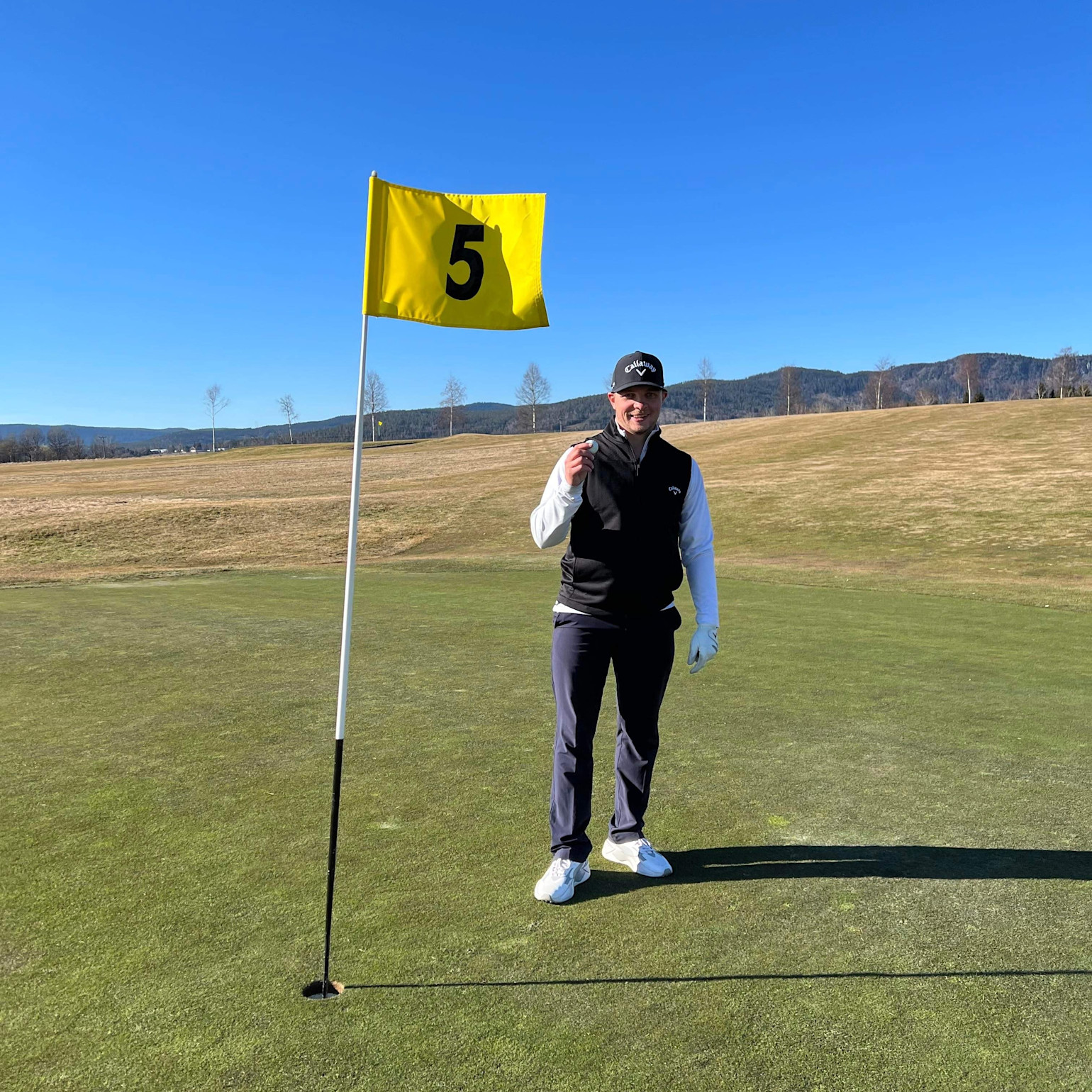 Man holder en golfbold ved siden af hul 5 på en golfbane under en klar blå himmel og omgivet af landskab.