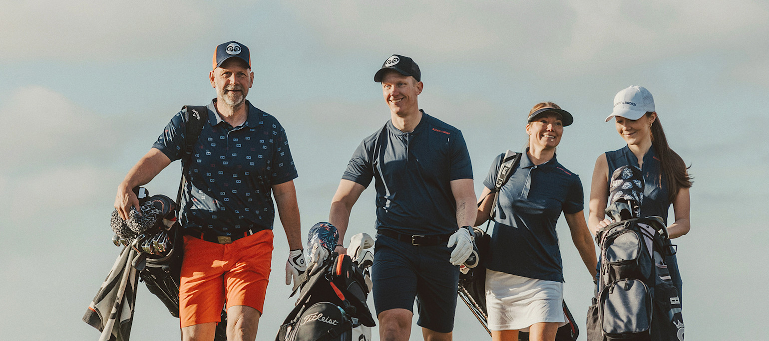 Fire personer går sammen på en golfbane, bærende på golfbags og iført golfbeklædning under en klar himmel.