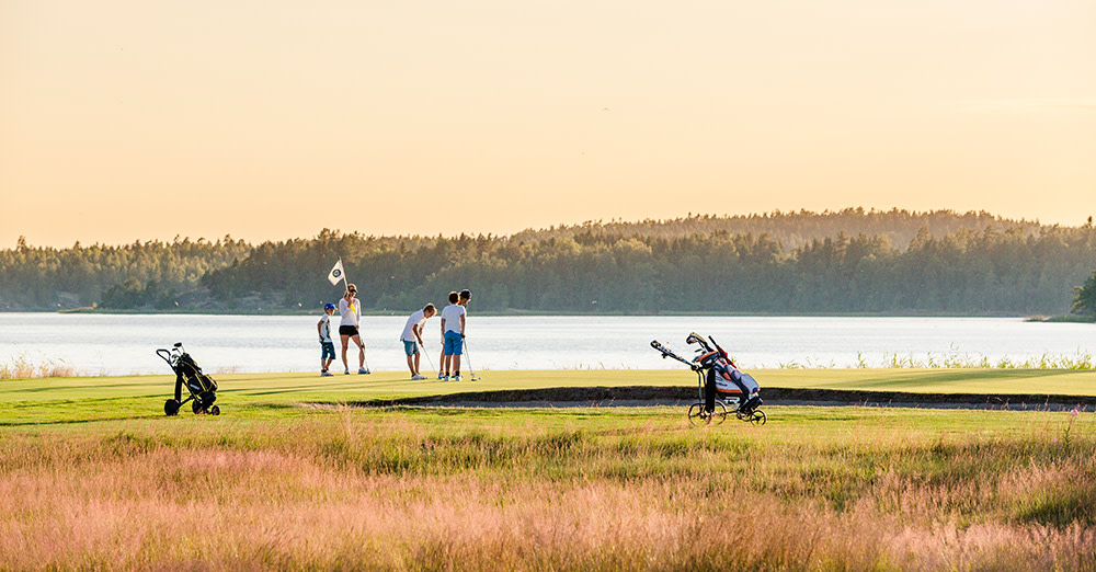 Flera personer spelar golf vid en green nära en sjö, med golfbagar på vagnar och skog i bakgrunden.