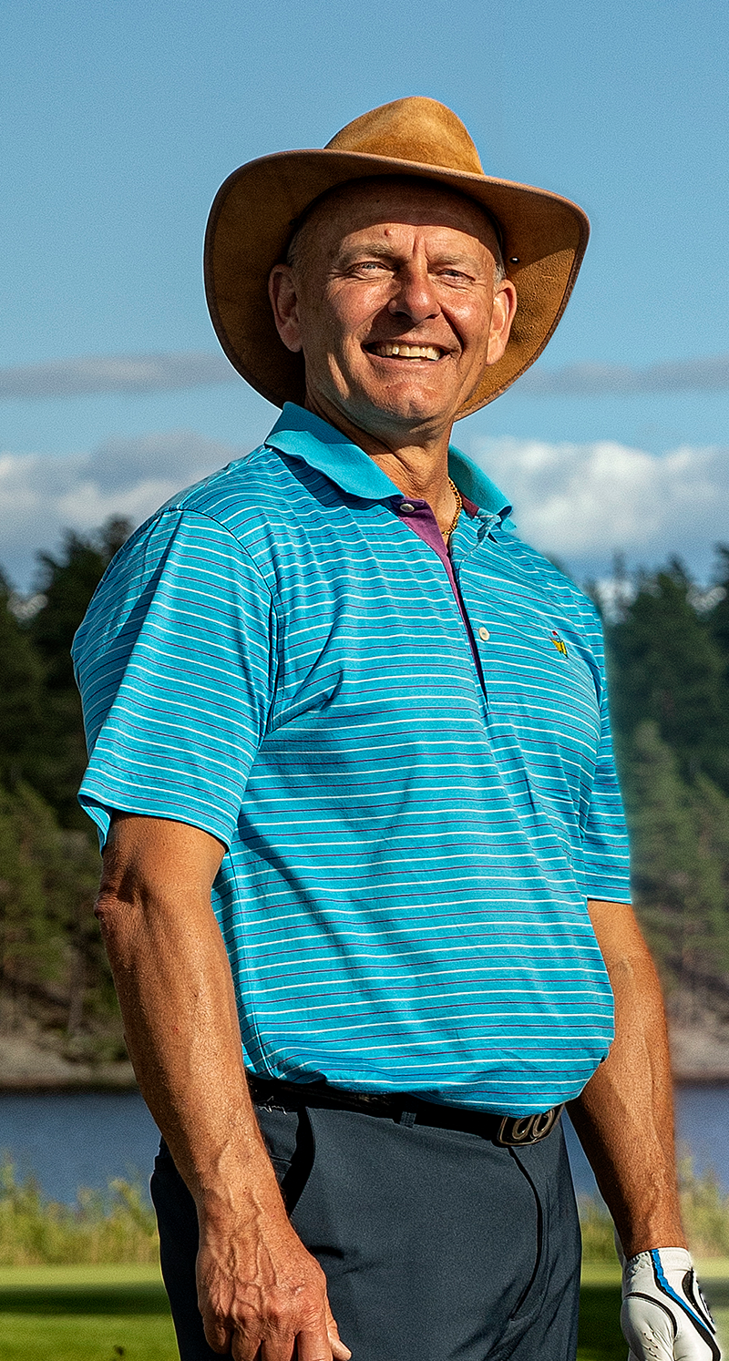 Man med cowboyhatt och blårandig pikétröja står utomhus på en golfbana med skog och vatten i bakgrunden.
