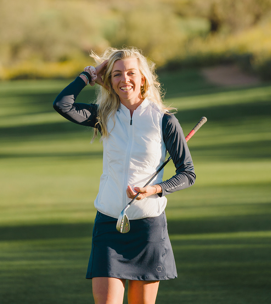 Kvinna med långt blont hår håller en golfklubba och ler på en solig golfbana.