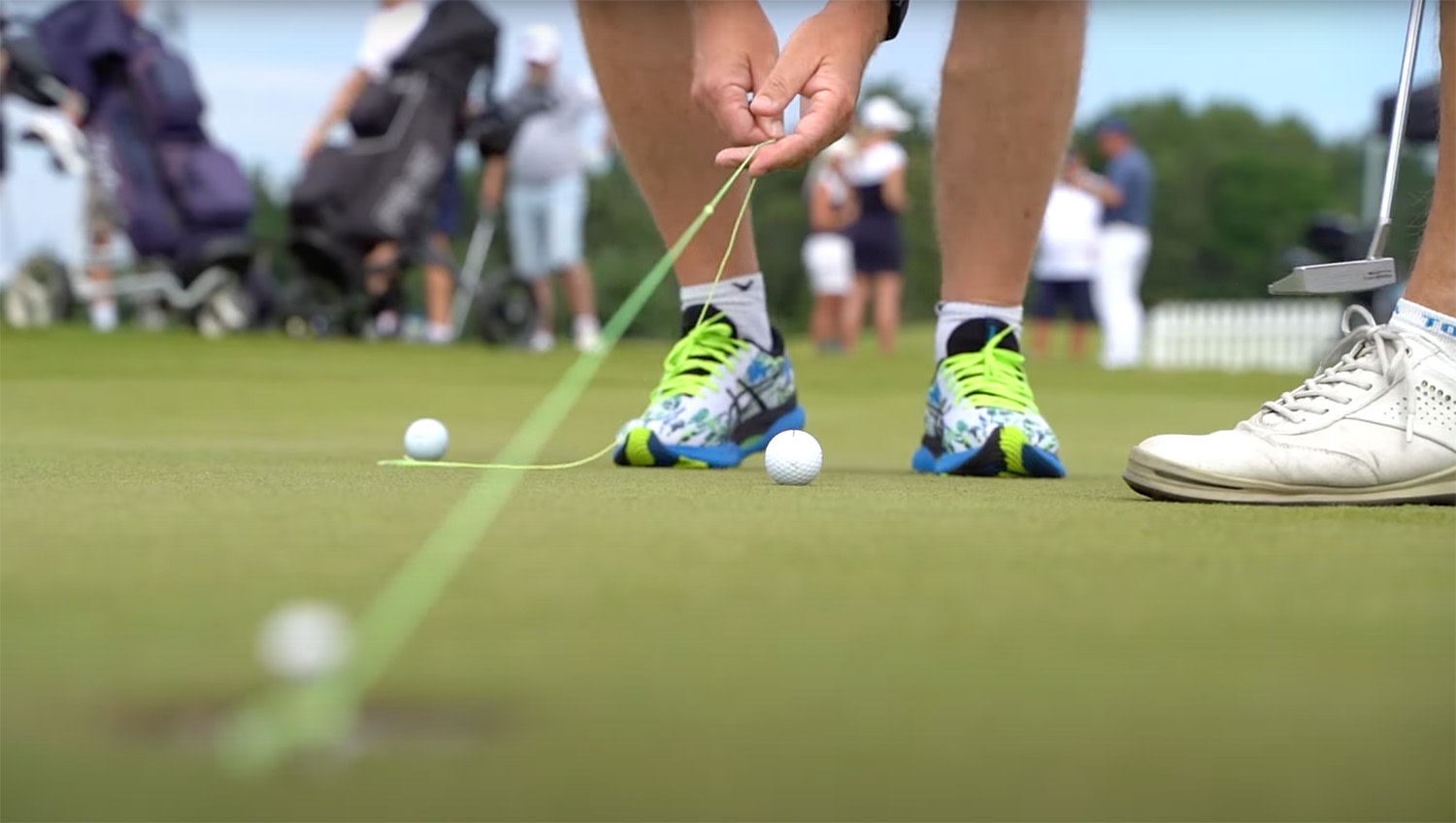 En person justerar ett snöre framför en golfboll på greenen, med golfklubba och golfskor, andra spelare syns i bakgrunden.