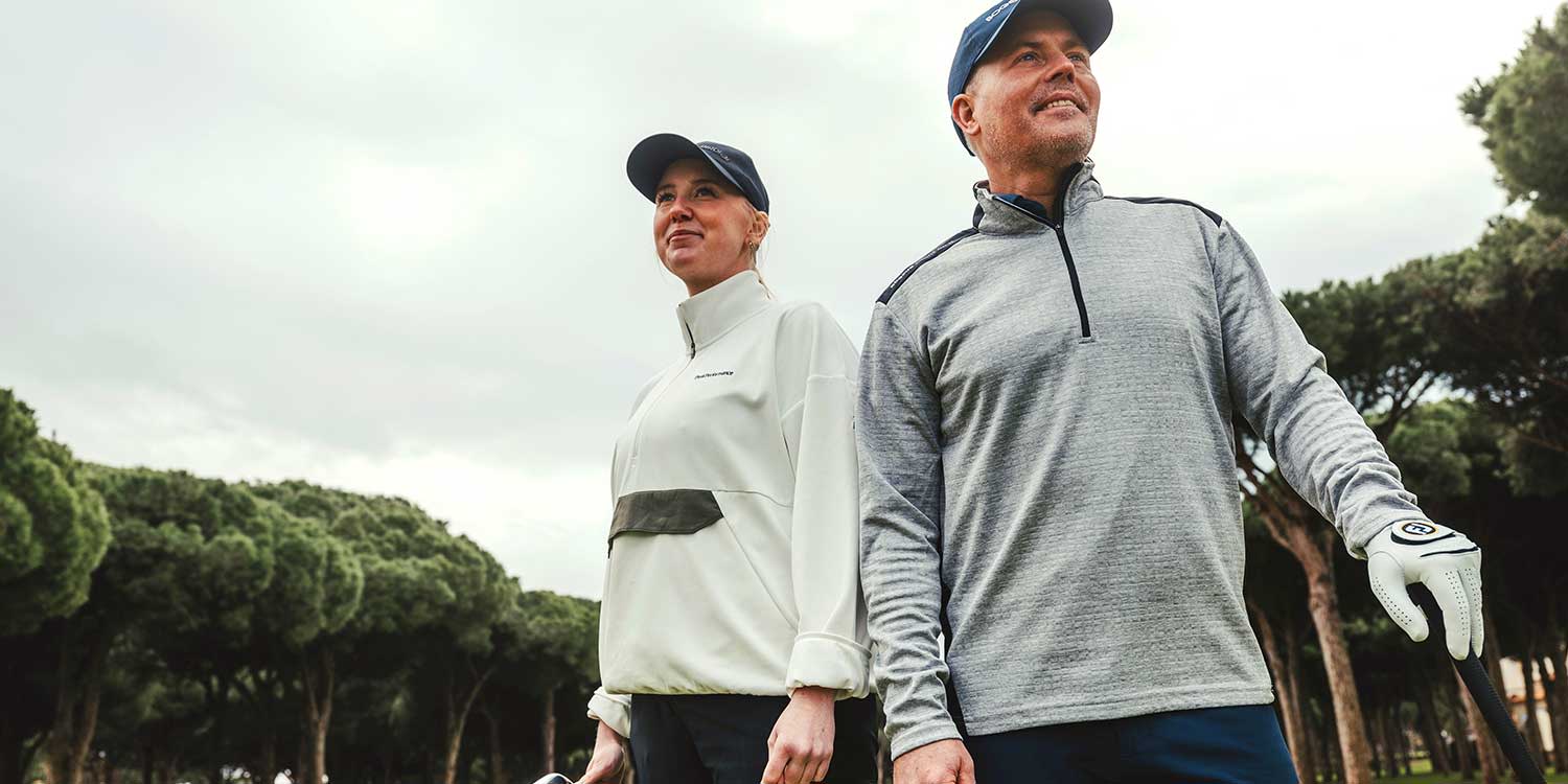 Two golfers are standing outdoors on a golf course, dressed in sportswear and caps, surrounded by green trees.