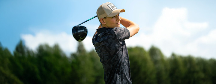Man i vit tröja och blå keps svingar en golfklubba utomhus med träd i bakgrunden.