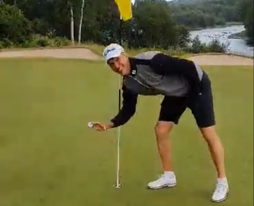 En mand iført golfbeklædning bøjer sig ned for at samle en golfbold op fra hullet på en golfbane med flag.