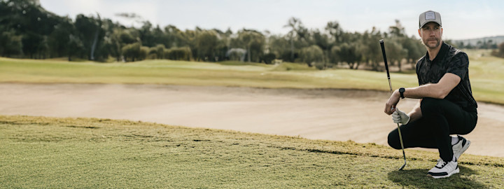 Man bär en golfbag över axeln och går utomhus på en golfbana, klädd i blå pikétröja och keps.