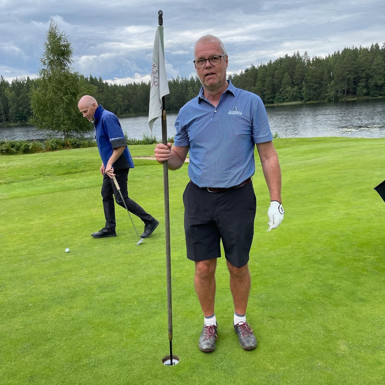 Man håller i en flaggstång vid ett golfhål på green, en annan man går bakom, sjö och skog i bakgrunden.