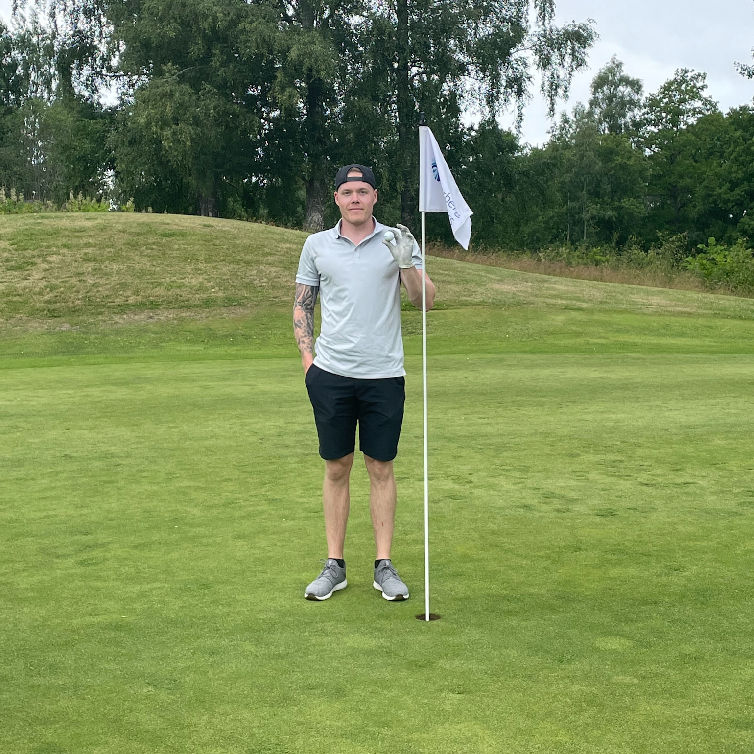 Mand i golfbeklædning står ved siden af en flagstang på en golfgreen, holder en golfhandske og laver en gestus med hånden. Skov i baggrunden.