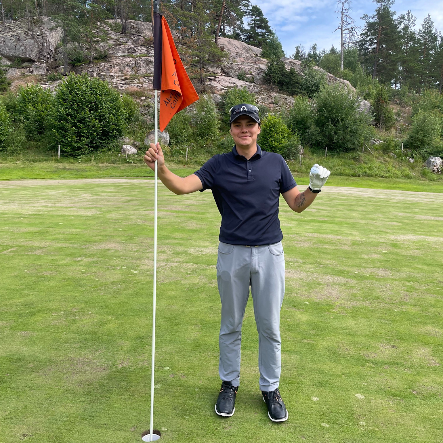 Person står på en golfgreen, håller i flaggstången med orange flagga och ser glad ut, omgiven av grönska och klippor.