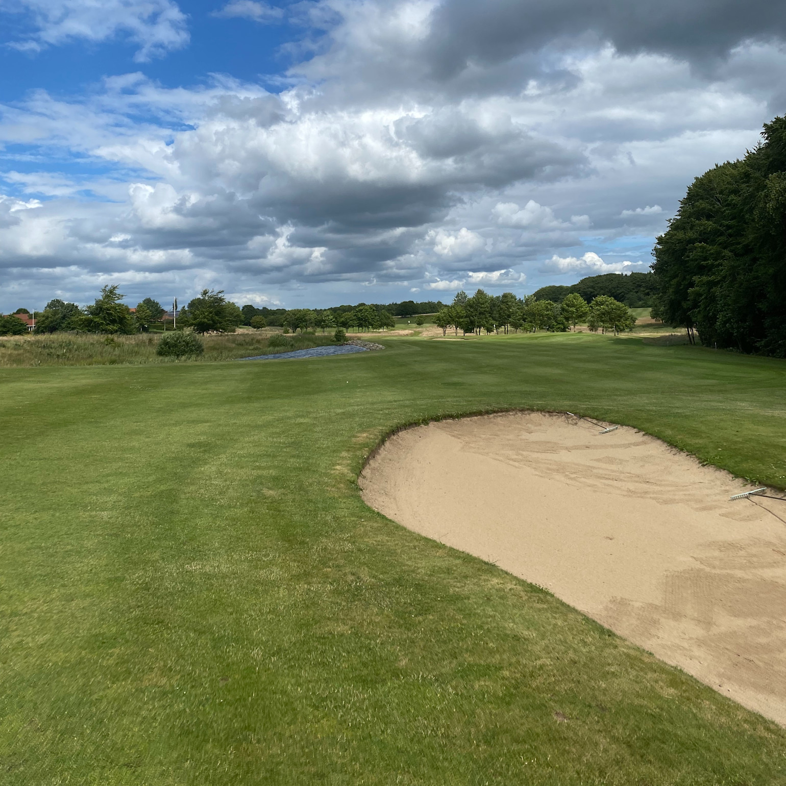 Sandbunker ligger på en golfbane med grønt græs, træer i baggrunden og overskyet himmel ovenover.