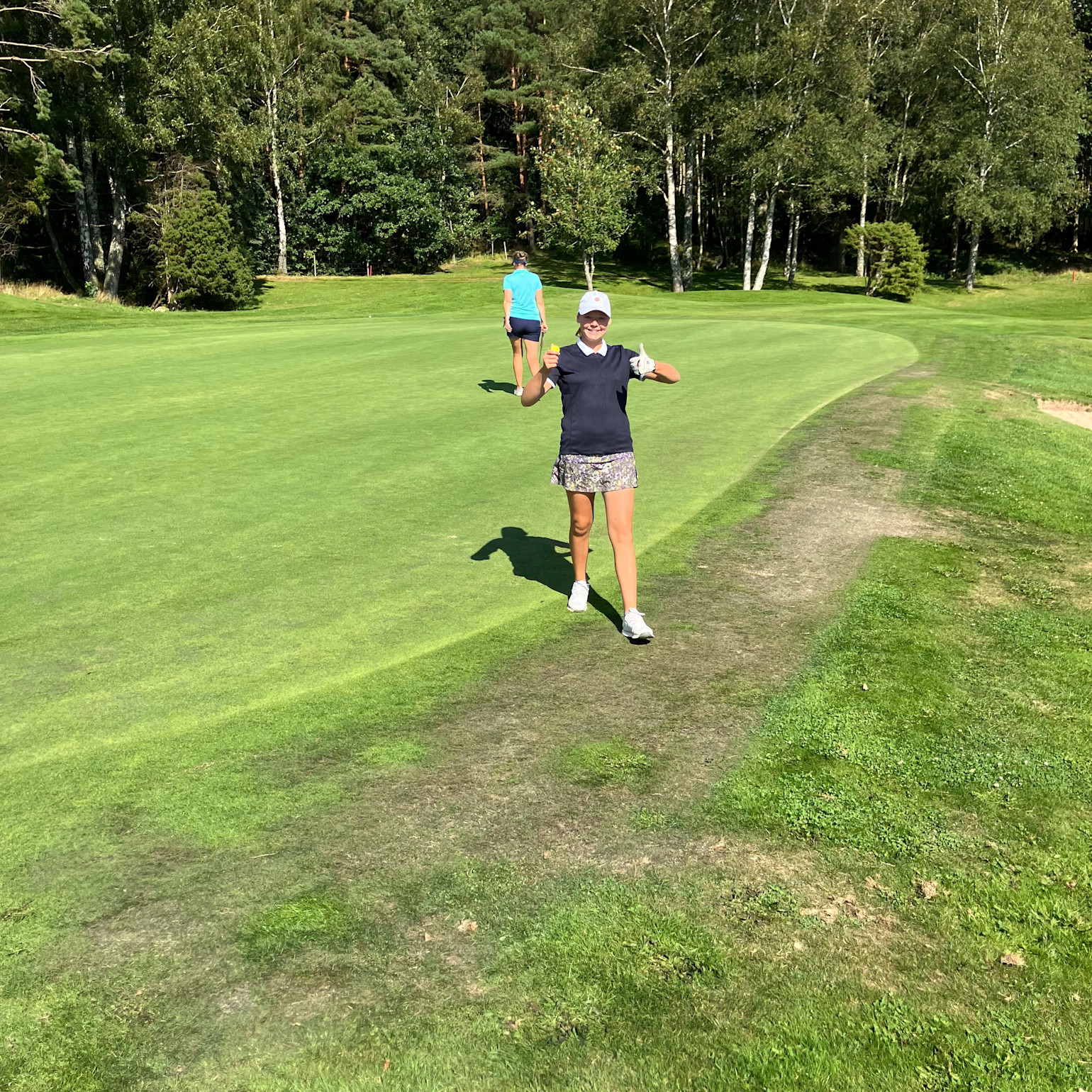 Kvinna i golfkläder ger tummen upp och ler på en golfbana, med en annan person längre bort och träd i bakgrunden.