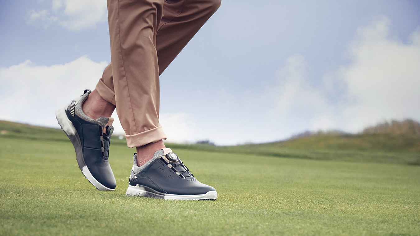 Person går på gräs i beige byxor och svarta sneakers med vit sula och grå detaljer under en blå himmel.