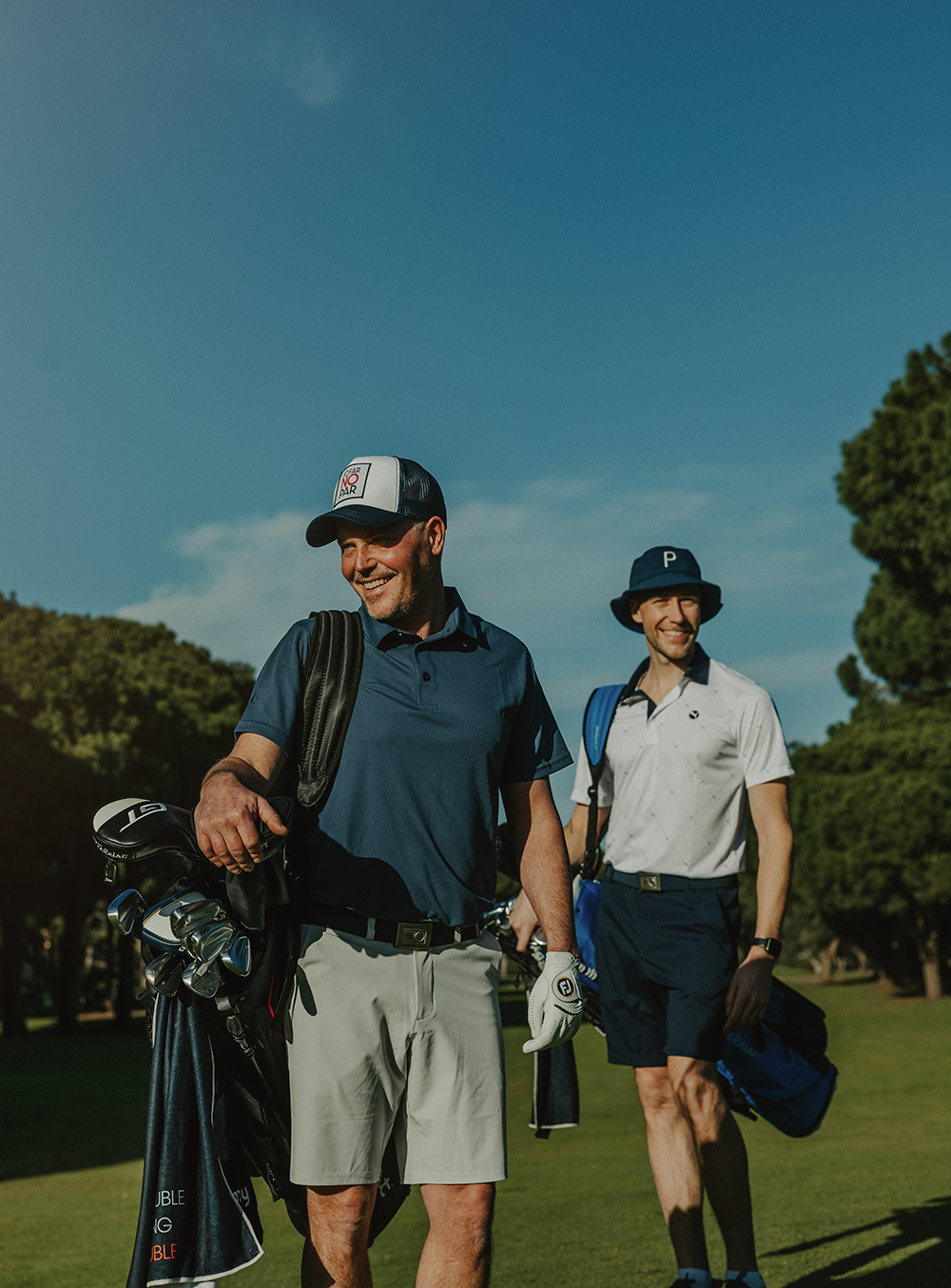 Kaksi miestä kantaa golfbägejä ja hymyilee kävellessään golfkentällä kirkkaan sinisen taivaan alla.