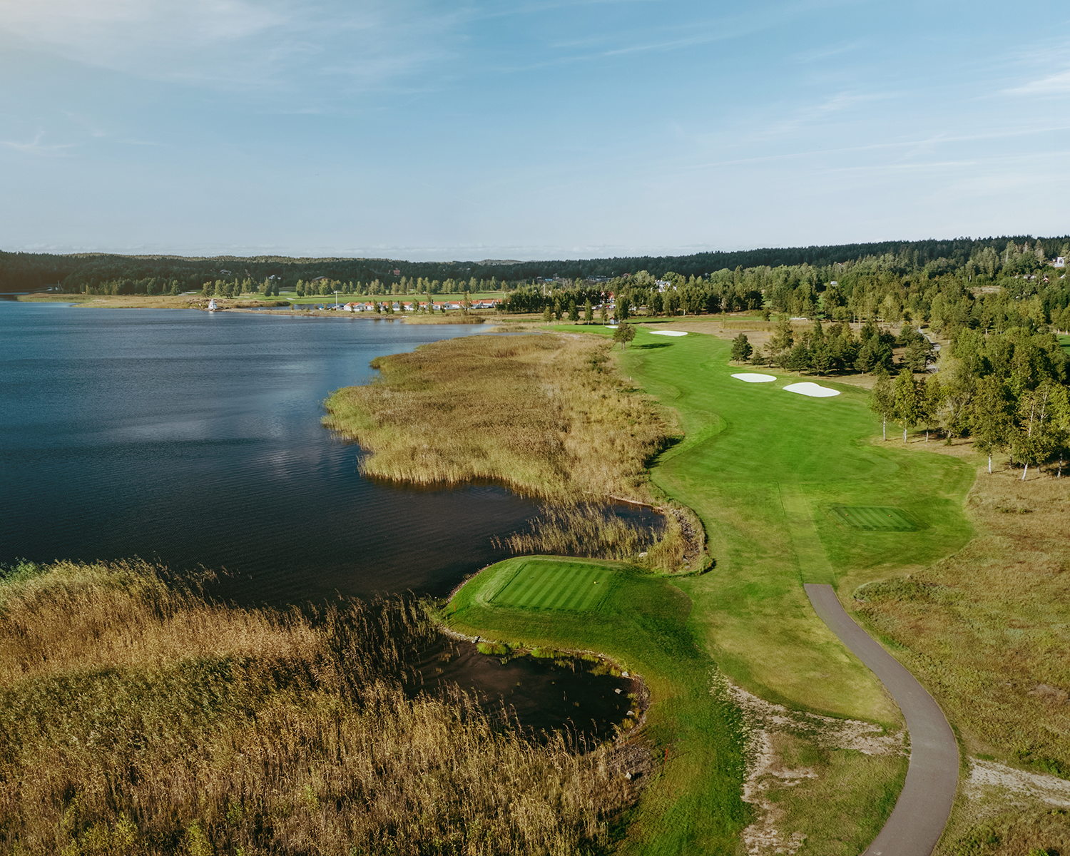 Golfbana med tee och fairway vid en sjö, omgiven av vass och träd under en klarblå himmel.