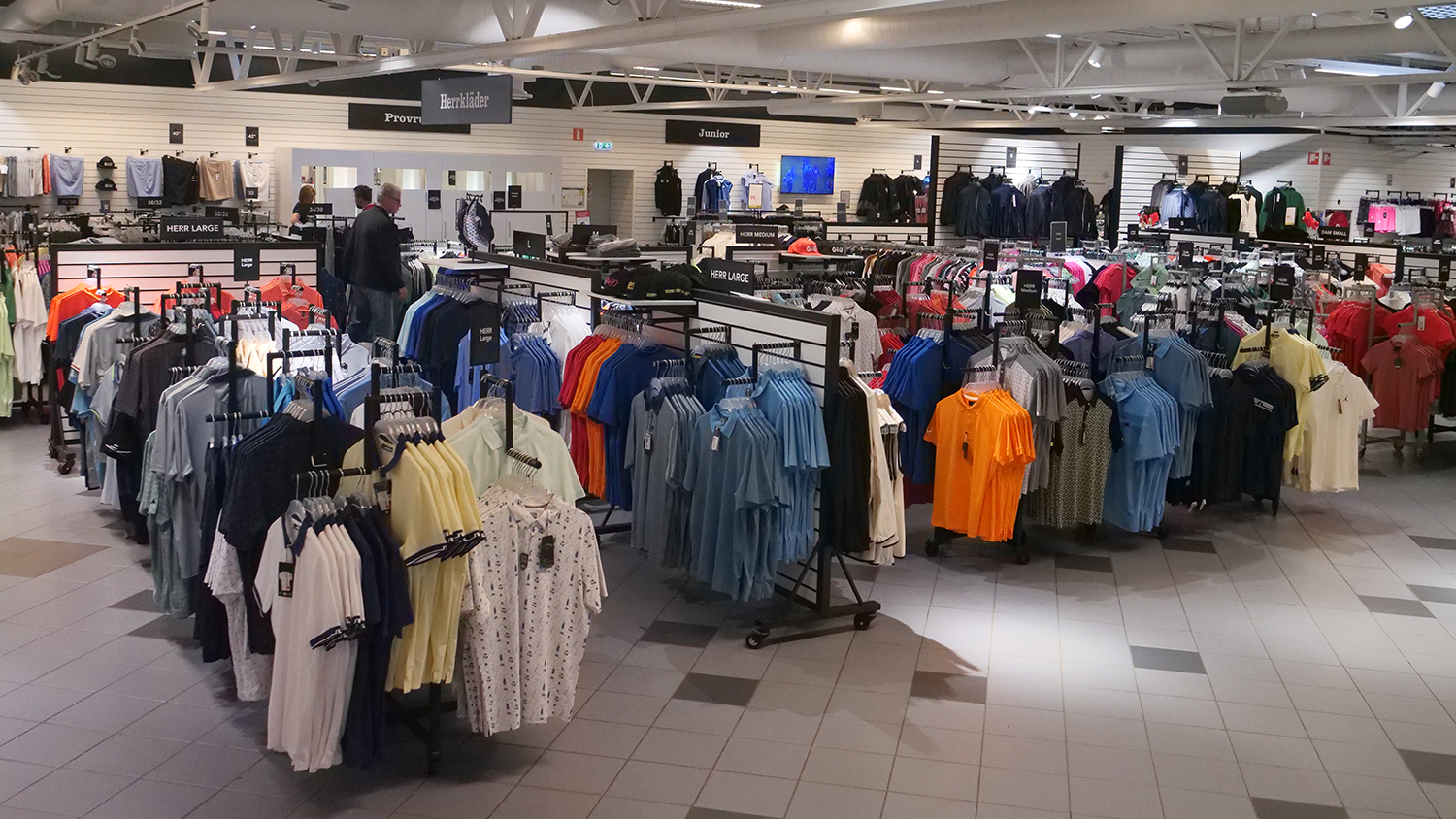 Klädställ med färgglada herrtröjor och skjortor i en butik, skyltar med storlekar och avdelningar syns i bakgrunden.