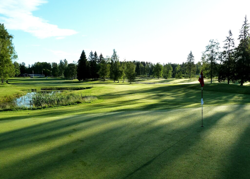 Golfhål med flagga på green, omgiven av gräs, träd och en liten damm under klar himmel.