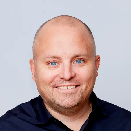 En leende skallig man med blå ögon och mörk skjorta mot en ljus bakgrund.