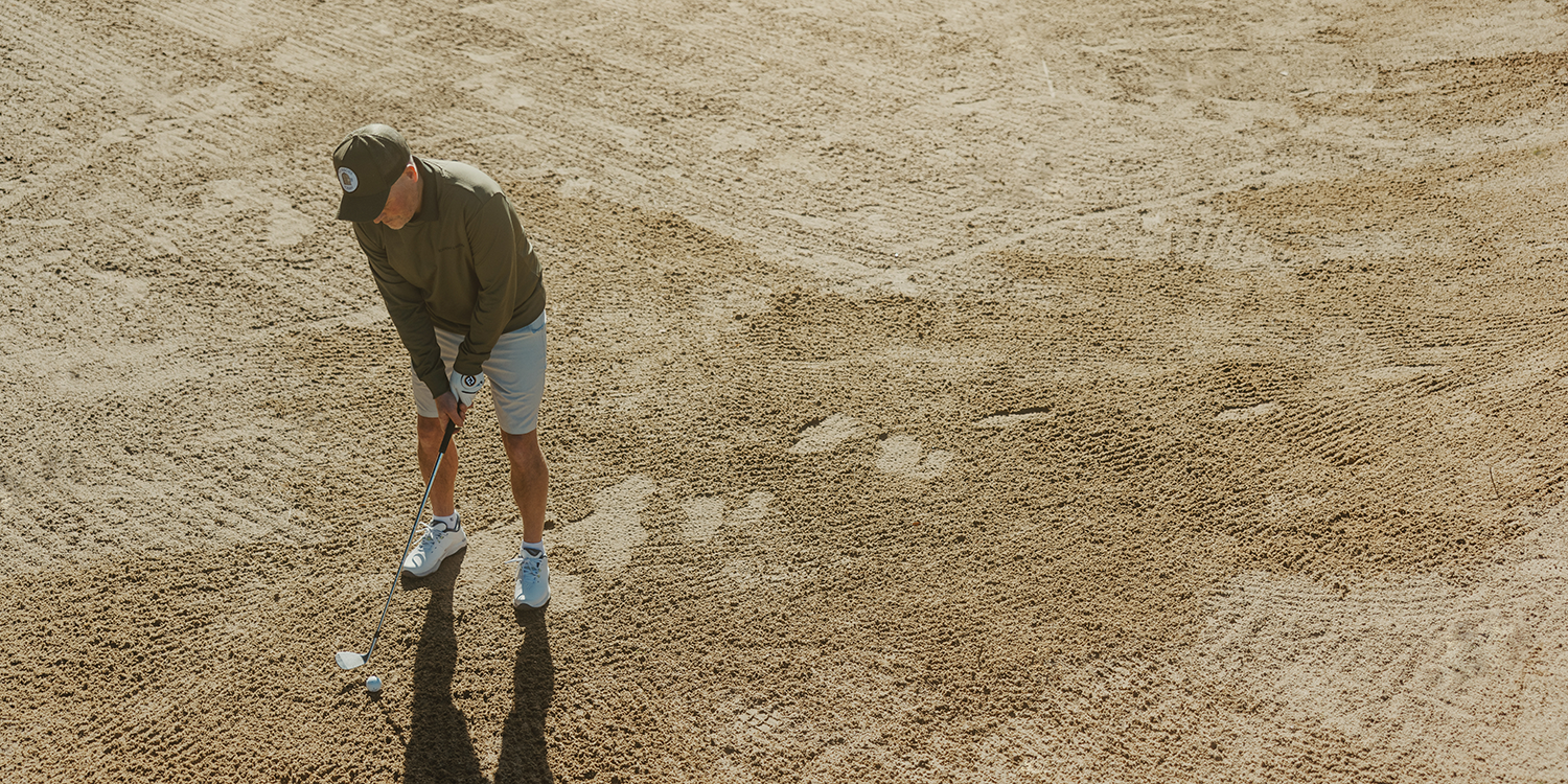 Ein Mann mit einer Kappe und Sportkleidung schlägt mit einem Golfschläger einen Golfball aus einem Sandbunker auf einem Golfplatz.