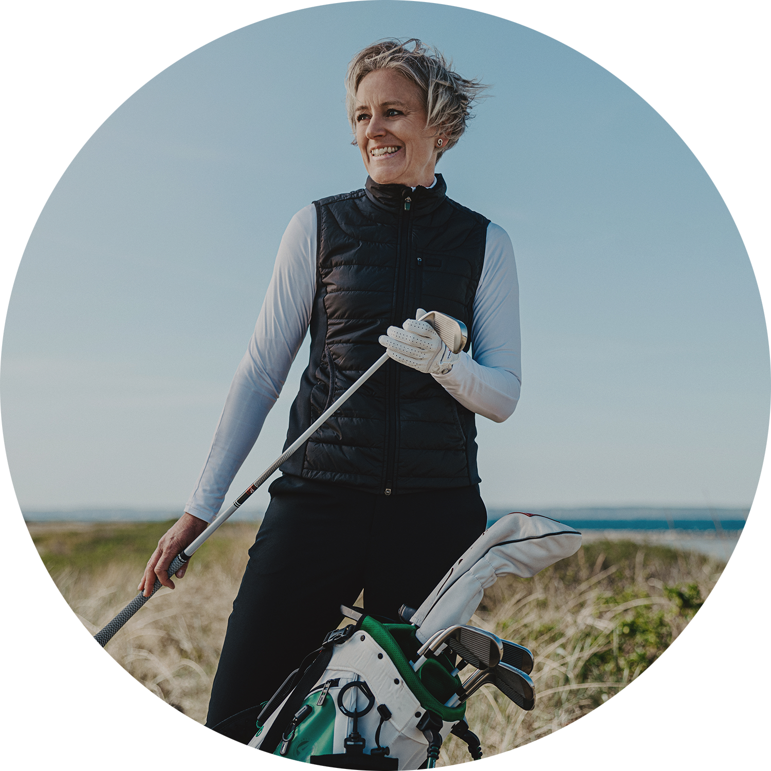 Nainen, jolla on lyhyet hiukset, pitää kädessään golfmailaa ja seisoo golfbägin vieressä ulkona golfkentällä.