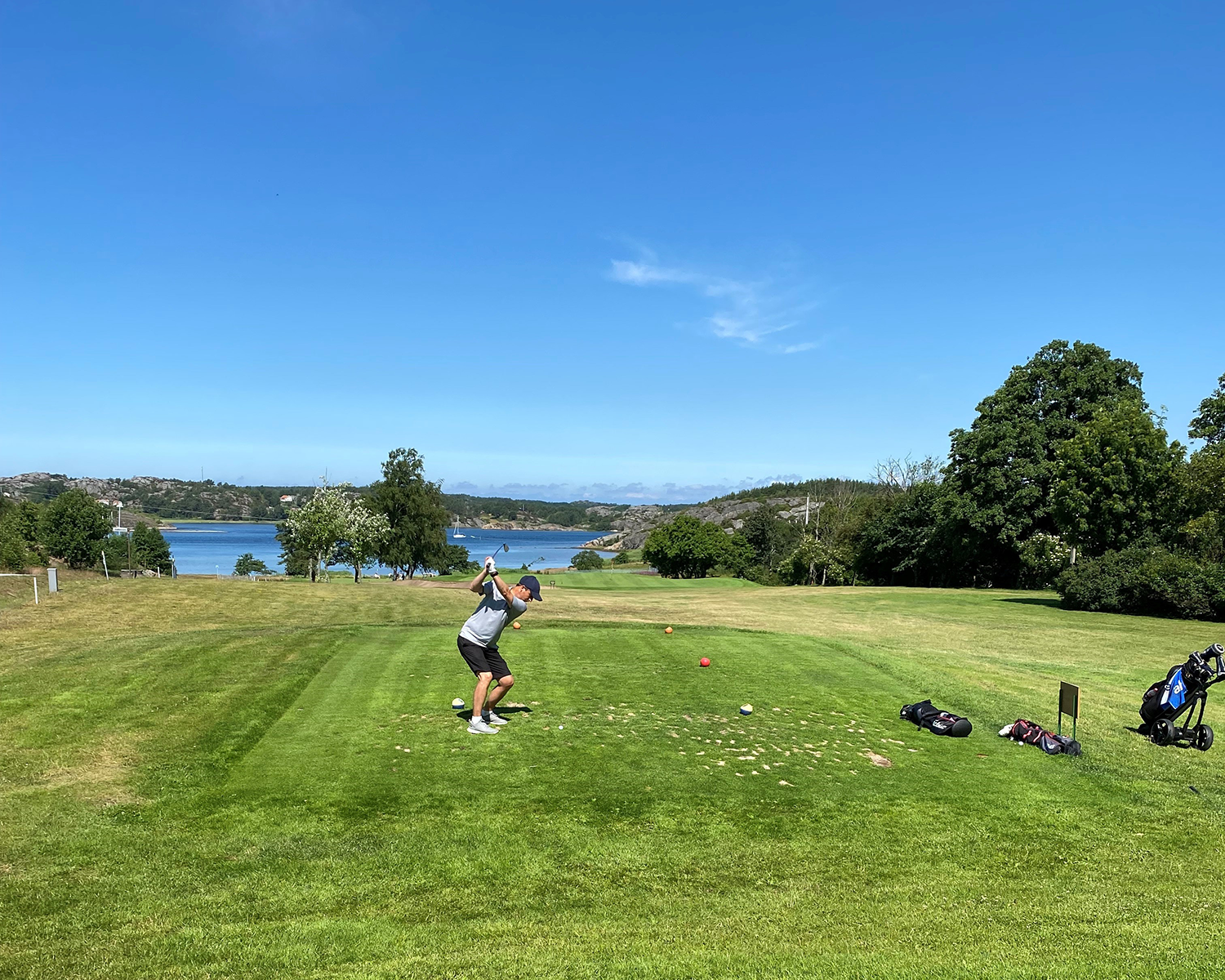 En golfspelare svingar en klubba på en grön golfbana med havsutsikt, omgiven av träd och blå himmel.