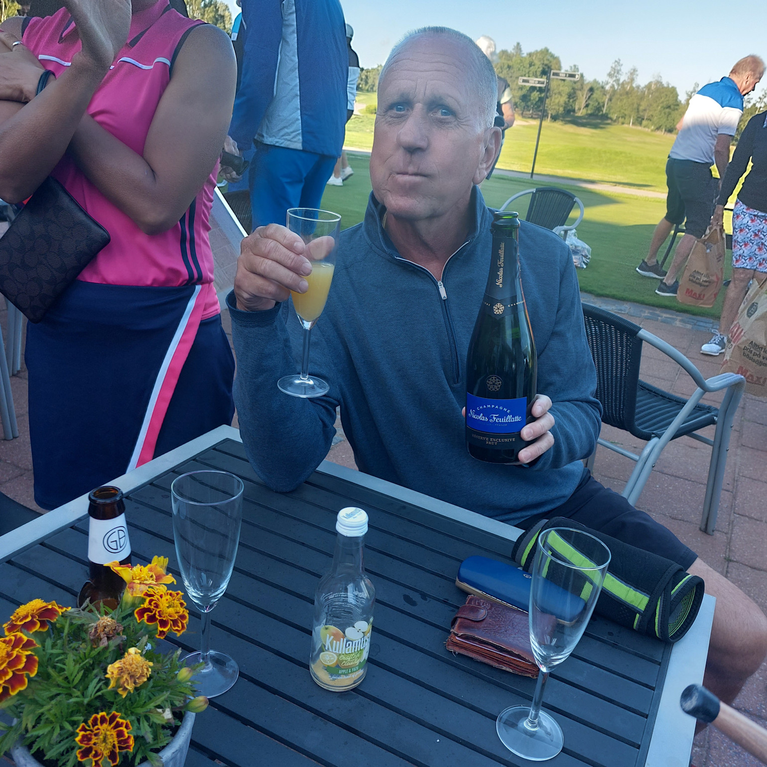 Mand i blå trøje sidder ved et bord udendørs, holder et champagneglas og en flaske Nicolas Feuillatte, omgivet af glas og drikkevarer.